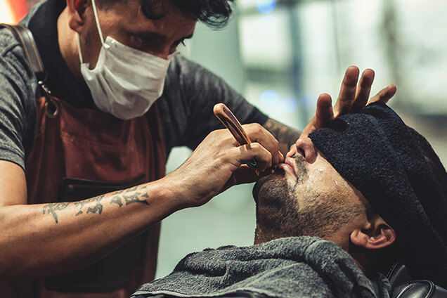 a barber with face mask on styling a beards of a man whose eyes have covered