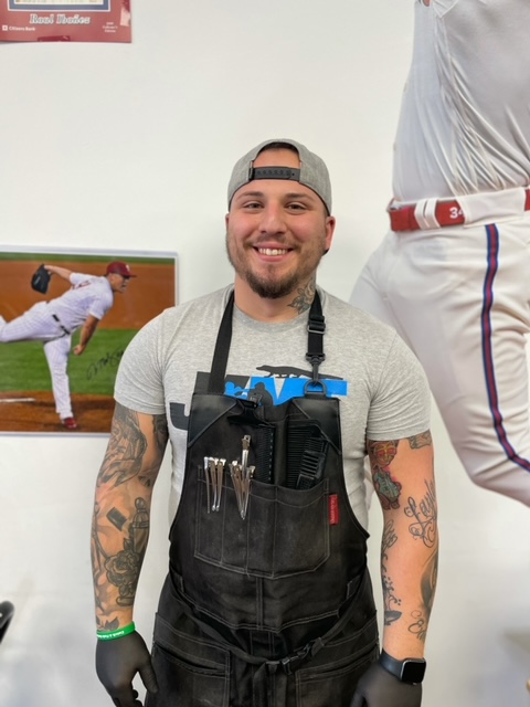A smiling barber in a salon, wearing an apron with scissors, tattoos on arms, baseball picture in background.