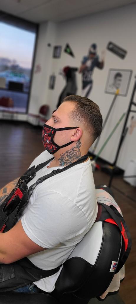 a man with tattoo on neck sitting in a salon wearing apron and face-mask after getting a haircut